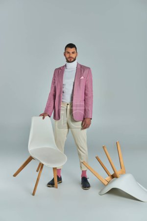 full length of man in lilac blazer and white pants posing with armchairs on grey, fashion and style