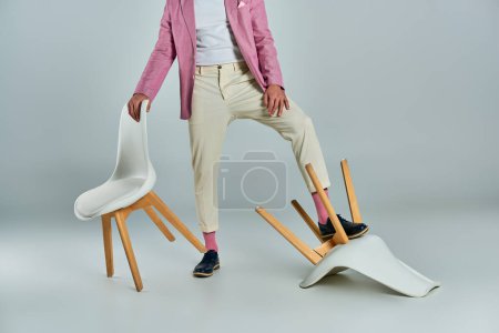 vue recadrée de l'homme en blazer lilas et pantalon blanc posant avec des fauteuils sur gris, business fashion