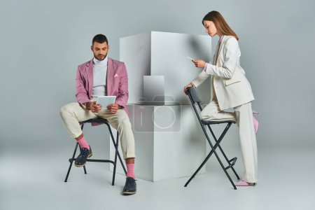 Stilvolles Paar mit Smartphone und digitalem Tablet in Laptopnähe auf weißen Würfeln vor grauem Hintergrund