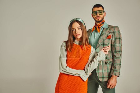 young and stylish couple in orange dress and plaid jacket looking at camera on grey, vintage fashion
