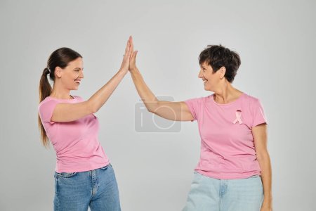 breast cancer concept, happy women with pink ribbons giving high five on grey backdrop, cancer free