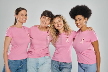 breast cancer awareness, cheerful interracial women with pink ribbons hugging on grey, diversity