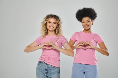 breast cancer awareness, happy multicultural women with pink ribbons on grey backdrop, heart sign
