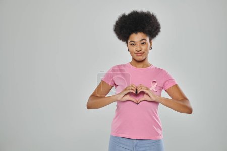 Téléchargez les photos : Sensibilisation au cancer du sein, femme afro-américaine avec ruban rose sur fond gris, signe cardiaque, sourire - en image libre de droit