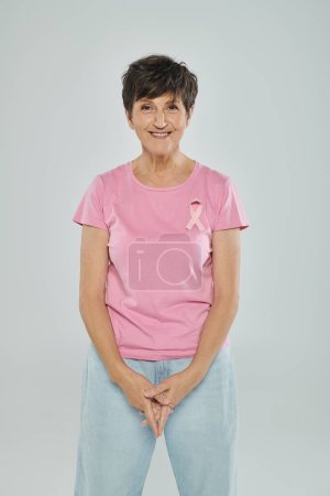 breast cancer awareness, happy middle aged woman with pink ribbon, grey backdrop, cancer free