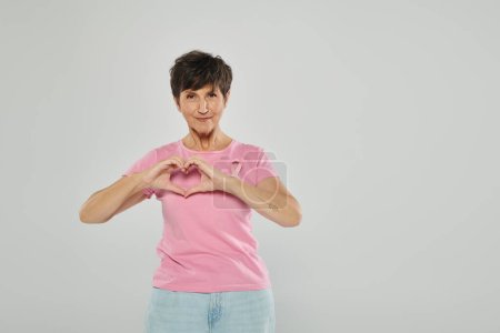 breast cancer awareness, happy mature woman with pink ribbon, grey backdrop, portrait, heart sign