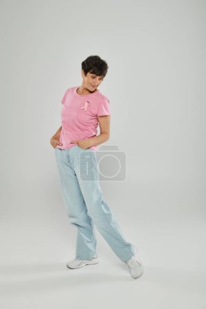 breast cancer awareness, happy mature woman with pink ribbon, grey backdrop, hands in pockets