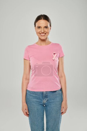 happy woman with pink ribbon, smiling, grey backdrop, breast cancer awareness, cancer free concept