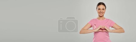 happy woman with pink ribbon, grey backdrop, breast cancer awareness, showing heart sign, banner