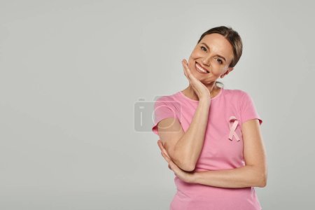 happy woman with pink ribbon on grey backdrop, breast cancer awareness, cancer free, smile and joy