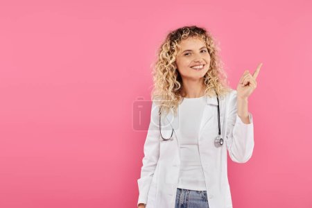 Brustkrebs-Bewusstseinskonzept, fröhliche Ärztin zeigt nach oben, rosa Hintergrund, Lächeln, Frau