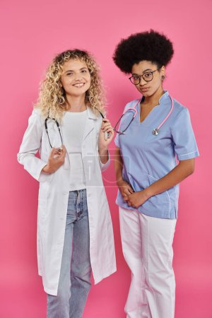 oncologists, interracial female doctors in white coats on pink backdrop, breast cancer awareness