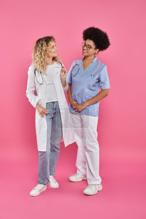 female oncologists, interracial doctors in white coats on pink backdrop, breast cancer awareness