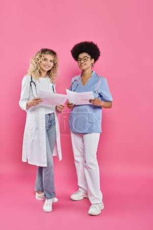 oncólogos femeninos alegres, doctores interracial en el telón de fondo rosado, conciencia del cáncer de pecho
