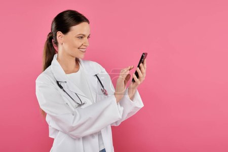 Lächelnde Ärztin im weißen Kittel mit Smartphone auf rosa, Brustkrebs-Konzept isoliert