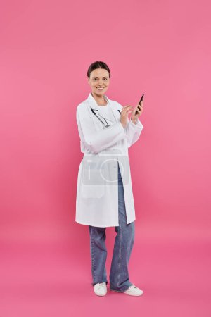 Téléchargez les photos : Médecin souriant en manteau blanc tenant smartphone et regardant la caméra sur rose, concept de cancer du sein - en image libre de droit