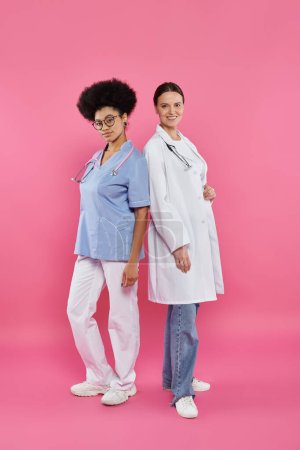 cheerful interracial doctors looking at camera on pink, breast cancer awareness concept