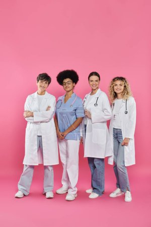full length of smiling interracial doctors standing together on pink, breast cancer month concept