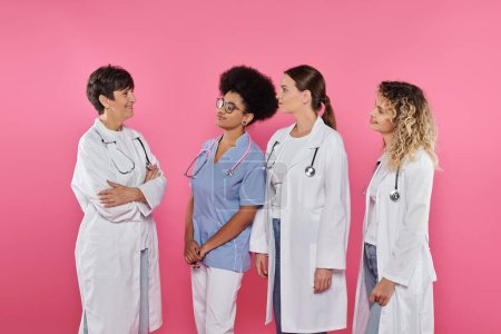 médicos multiétnicos mirando sonriente colega cruzando brazos aislados en rosa, cáncer de mama mes