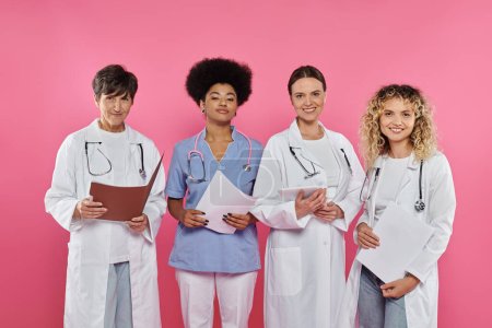 Foto de Médicos multiétnicos sonrientes sosteniendo carpetas de papel y tableta digital aislados en rosa, cáncer de mama - Imagen libre de derechos