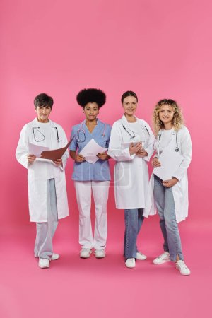 Foto de Sonrientes médicos interraciales sosteniendo carpetas de papel y tableta digital en rosa, mes del cáncer de mama - Imagen libre de derechos