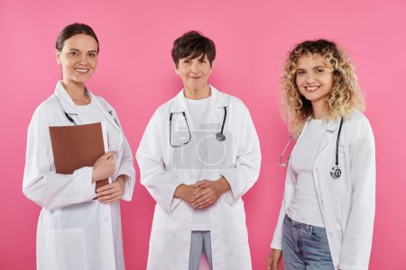 smiling female doctors in white coats looking at camera isolated on pink, breast cancer awareness