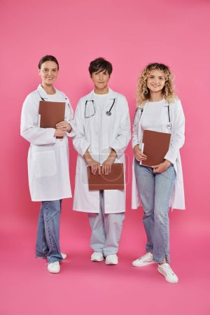 happy female doctors holding paper folders and standing on pink background, breast cancer awareness