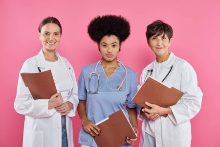 Téléchargez les photos : Médecins interracial souriants en manteaux blancs tenant des dossiers en papier isolés sur rose, cancer du sein - en image libre de droit