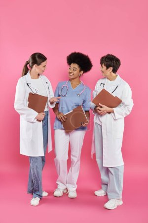 smiling multiethnic doctors with paper folders talking on pink, breast cancer awareness