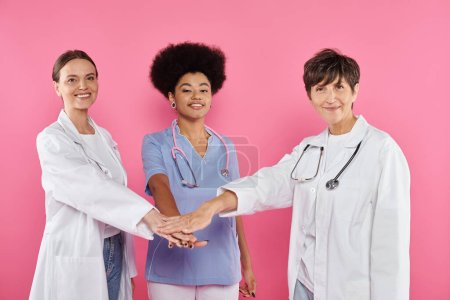 Foto de Oncólogos multiétnicos positivos tomados de las manos aislados en rosa, conciencia sobre el cáncer de mama - Imagen libre de derechos