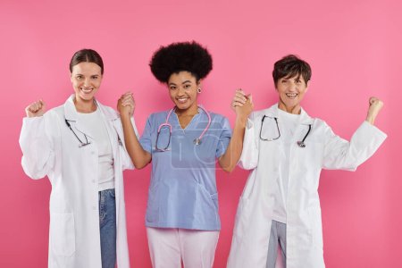 Foto de Médicos oncólogos multiétnicos emocionados tomados de las manos aislados en rosa, mes del cáncer de mama - Imagen libre de derechos