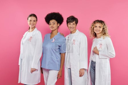 lächelnde interrassische Ärzte mit Schleifen, die in die Kamera schauen, isoliert auf rosa, Brustkrebs-Konzept