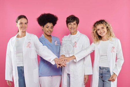 Foto de Médicos multiétnicos sonrientes con cintas agarradas de las manos aisladas en rosa, concepto de cáncer de mama - Imagen libre de derechos