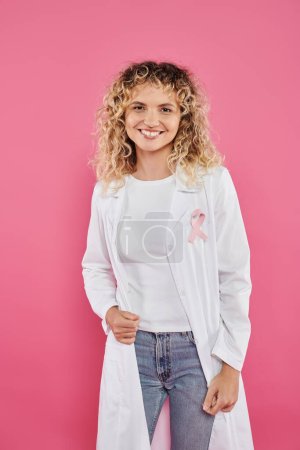 Photo for Smiling doctor with ribbon on white coat standing isolated on pink, breast cancer concept - Royalty Free Image