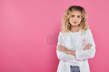 médecin bouclé avec ruban sur manteau blanc croisant les bras isolé sur rose, mois du cancer du sein