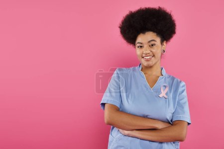 médecin afro-américain souriant avec ruban traversant les bras isolé sur rose, concept de cancer du sein