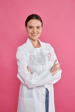 cheerful female oncologist with ribbon crossing arms isolated on pink, breast cancer month