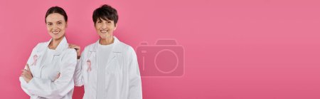 Photo for Smiling doctors with ribbons on white coats posing isolated on pink, breast cancer concept, banner - Royalty Free Image