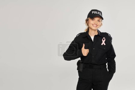 smiling policewoman with pink ribbon showing thumb up isolated on grey, breast cancer month