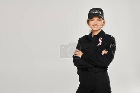 positive policewoman with pink ribbon crossing arms isolated on grey, breast cancer concept