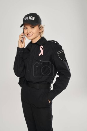 happy policewoman with pink ribbon on uniform talking on smartphone isolated on grey, breast cancer