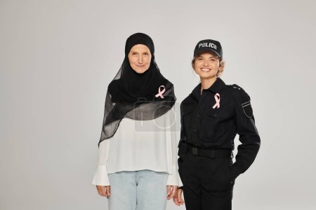 smiling policewoman and woman in hijab with pink ribbons of breast cancer isolated on grey