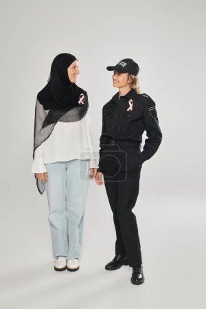 positive policewoman and woman in hijab with pink ribbons of breast cancer standing on grey