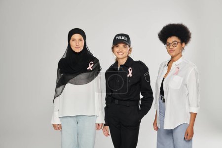 smiling policewoman, woman in hijab and african american woman with pink ribbons isolated on grey