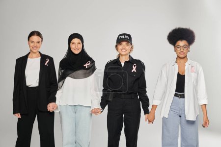 smiling different interracial women with pink ribbons holding hands isolated on grey, breast cancer