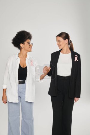 smiling businesswoman and african american woman with pink ribbons holding hands isolated on grey