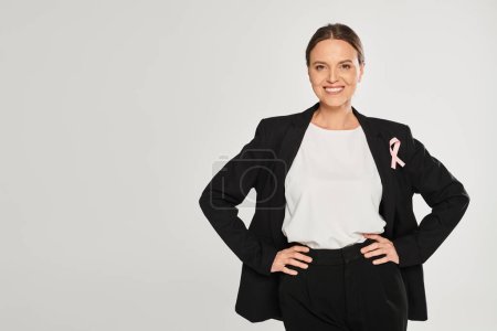 smiling businesswoman with pink ribbon on jacket posing at camera isolated on grey, breast cancer