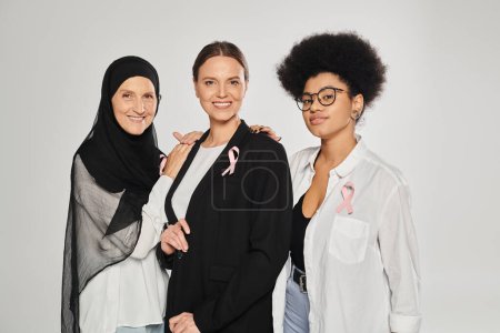 smiling interracial women with pink ribbons of breast cancer hugging isolated on grey