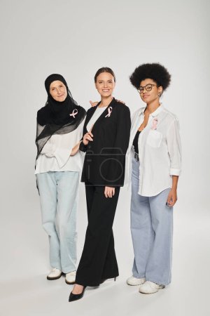 smiling interracial different women with pink ribbons of breast cancer standing on grey