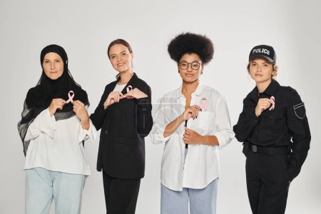smiling different and multiethnic women showing pink ribbons of breast cancer isolated on grey